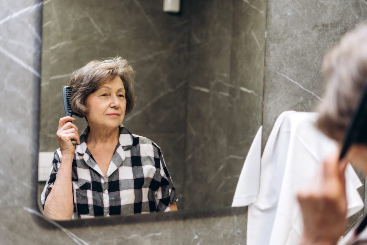 Vyresnio amžiaus moteris šukuoja plaukus prieš veidrodį kaip dalį atkuriamos priežiūros rutinos.