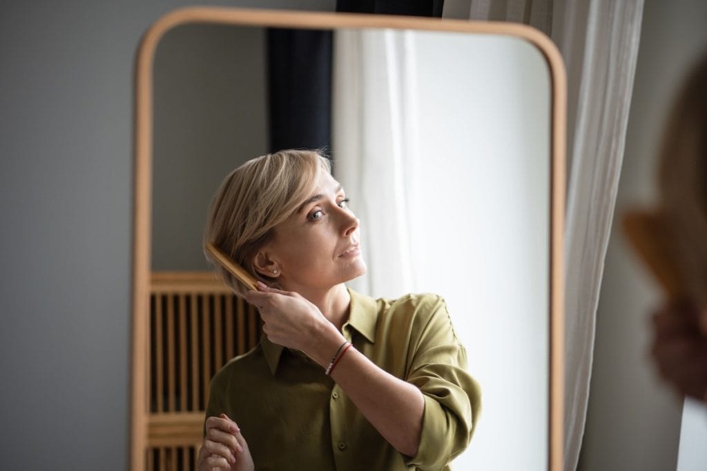 Moteris virš 40 metų tvarkosi plaukus prie veidrodžio, pabrėžiamas natūralus kirpimas