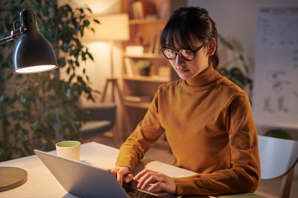 Stilingas biuras namuose, moteris dirba prie nešiojamojo kompiuterio su stalo lempa ir jaukia aplinka