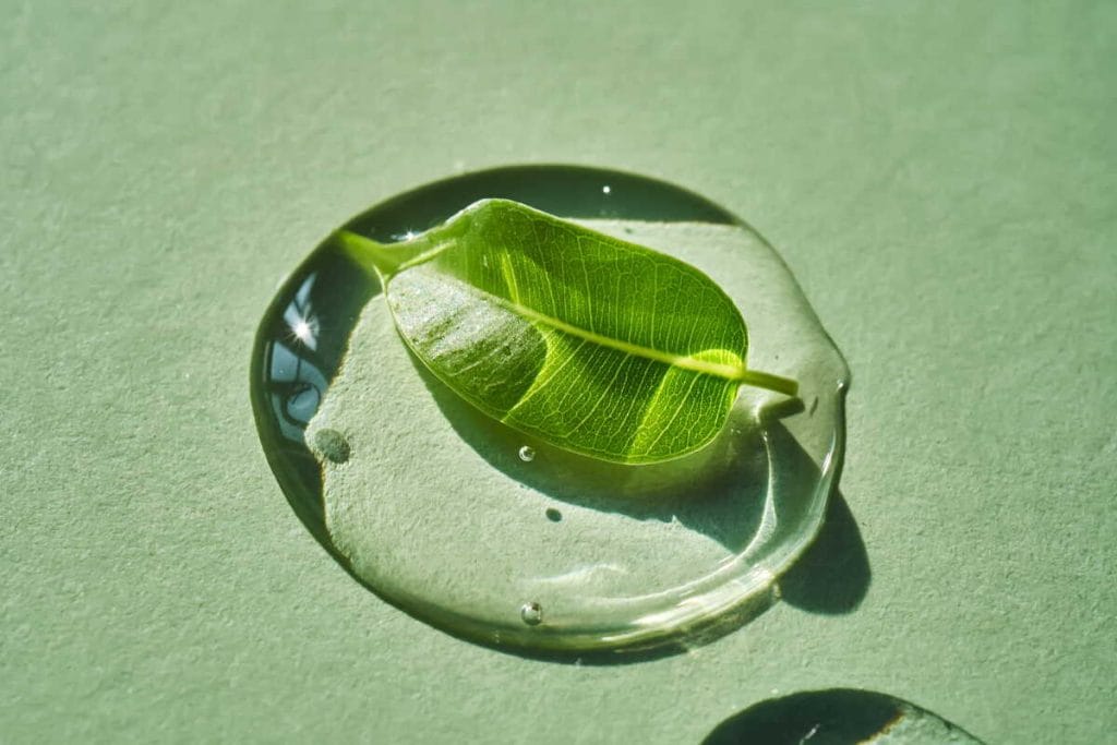 Žalias lapas guli skaidriame serumo laše ant šviesaus paviršiaus