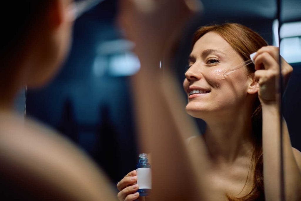 Moteris prieš veidrodį naudoja veido serumą – retinolio serumas dažnai įtraukiamas į vakarinę odos priežiūros rutiną