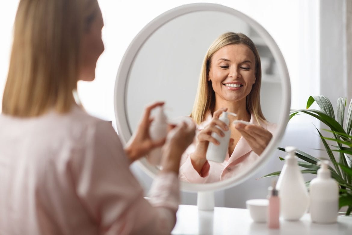 Moteris prie veidrodžio šypsodamasi tepa kremą ant švarios odos – tinkamai nuvalytas makiažas leidžia geriau įsisavinti priežiūros priemones.
